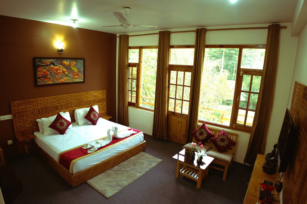 Une chambre d’hôtel avec un grand lit, une tête de lit en bois, un tableau, une fenêtre avec des rideaux, une petite table avec un vase et un banc en bois.