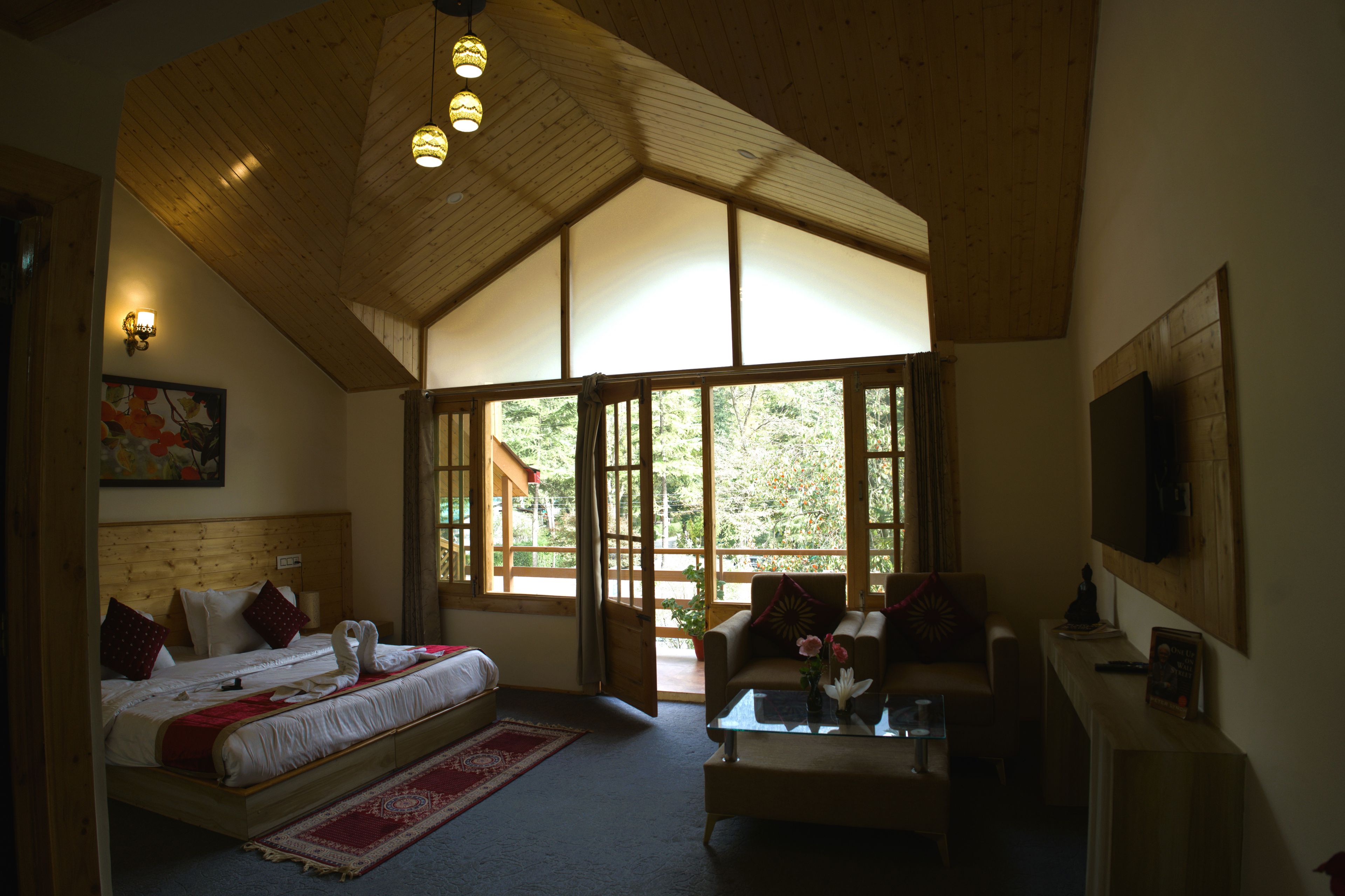 A bedroom with a large bed, a sofa, a coffee table, and a TV. There are windows and a wooden ceiling.