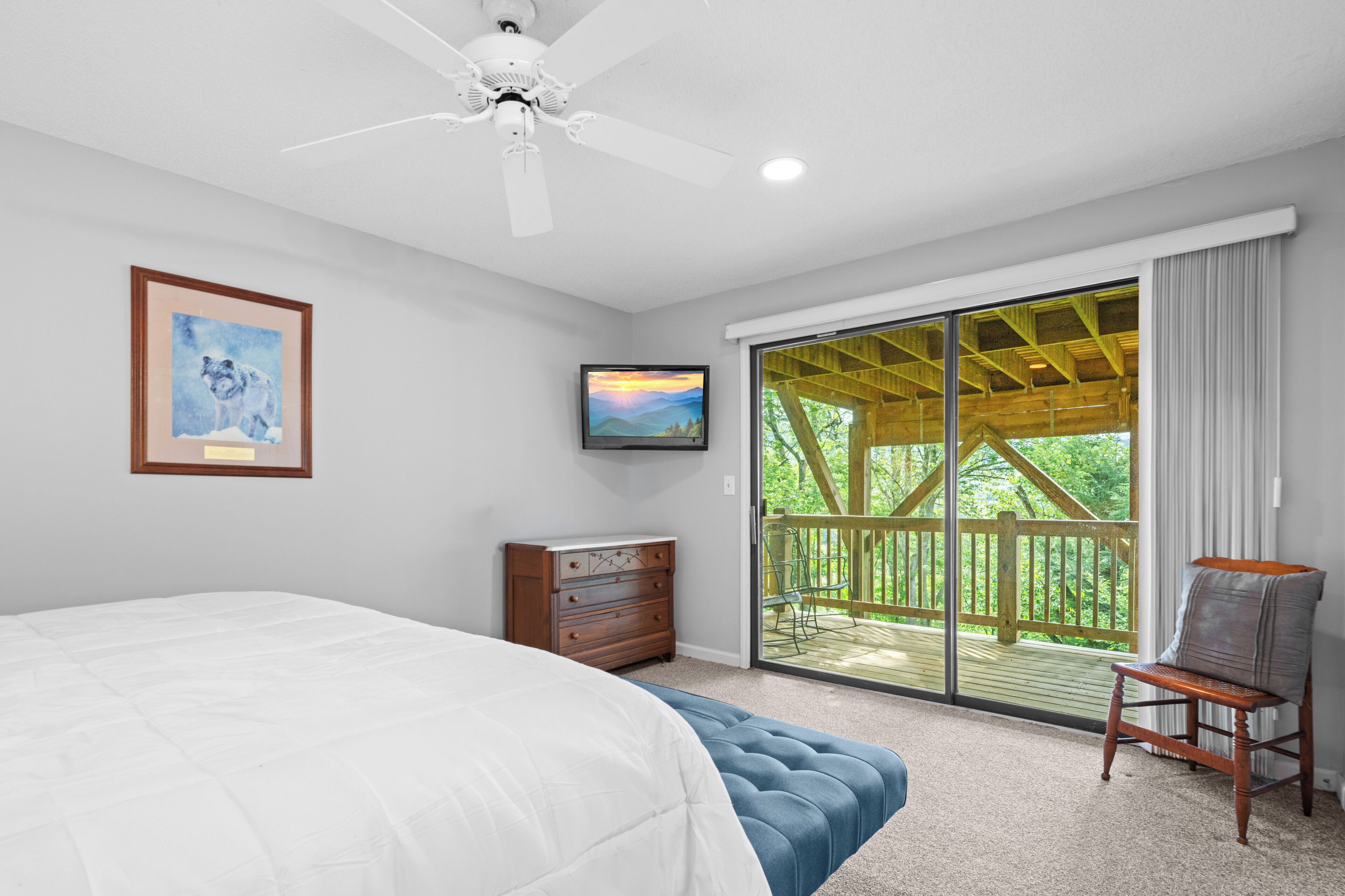 A bedroom with a large bed, a ceiling fan, a wooden dresser, a television, and a sliding glass door leading to a balcony with a wooden deck.