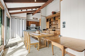 Ein Essbereich mit Holztisch und Stühlen, eine Küche im Hintergrund und ein großes Fenster mit Vorhängen.