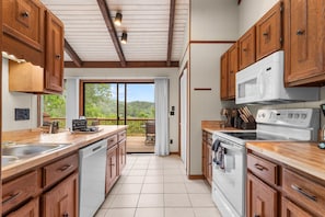 A kitchen with wooden cabinets, a white microwave, a stove, and a dishwasher. There is a sliding glass door leading to a balcony with a view of greenery. - Spacious 5BR Sugar Mt Cabin w/ Hot Tub & Views! (Sugar Mountain)