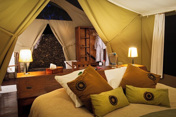 A tented accommodation with a bed, pillows, a bedside table, a lamp, a wooden chair, a small table, and a storage box. - Karen Blixen Camp (Narok)