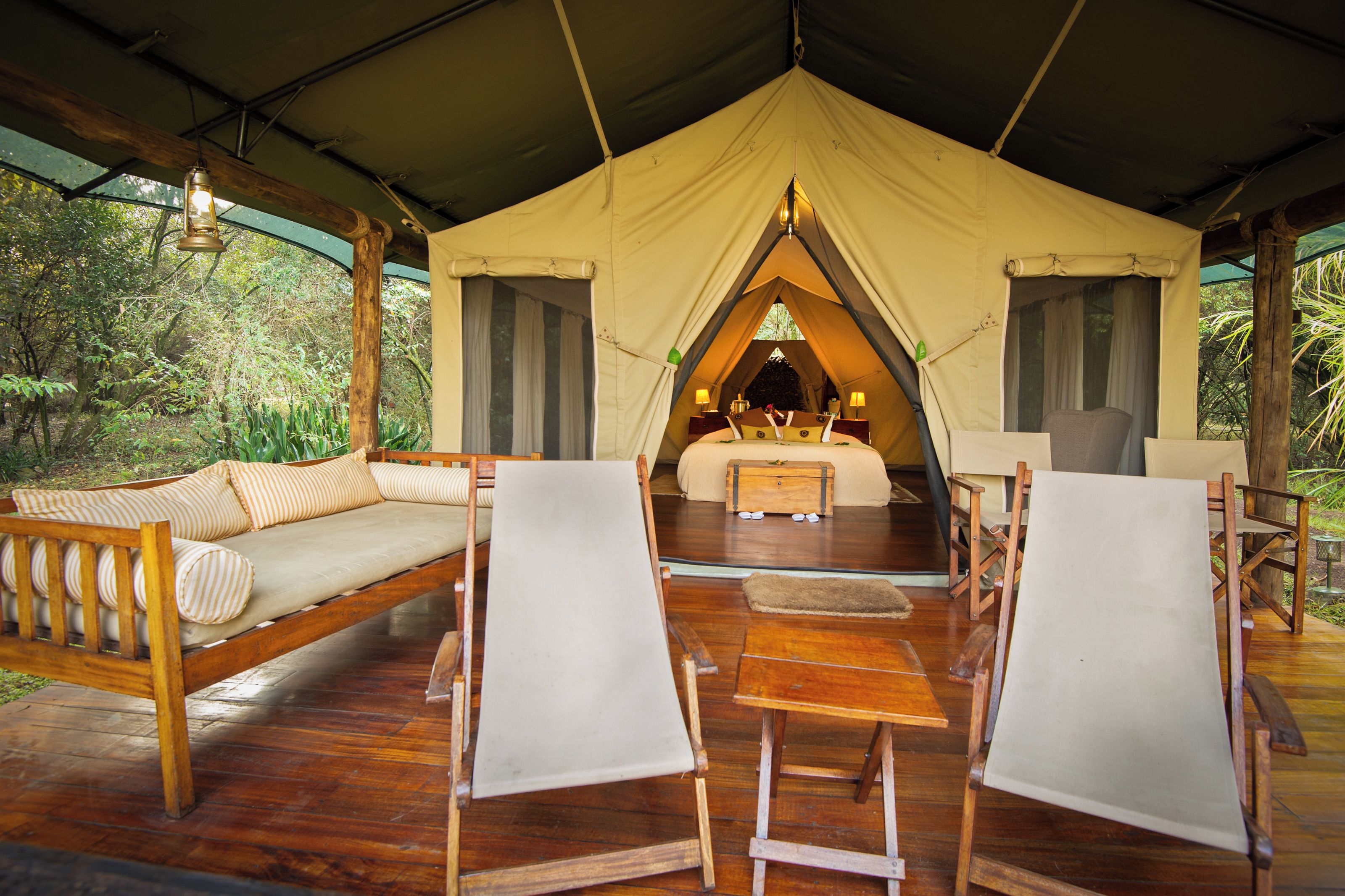 A tented accommodation with a wooden deck, a sofa, two chairs, a table, and a bed with a canopy.