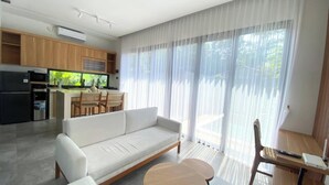 A modern living room with a sofa, a dining area with chairs, a kitchen with appliances, and large windows with sheer curtains. - Terra House B1 (Kedonganan)