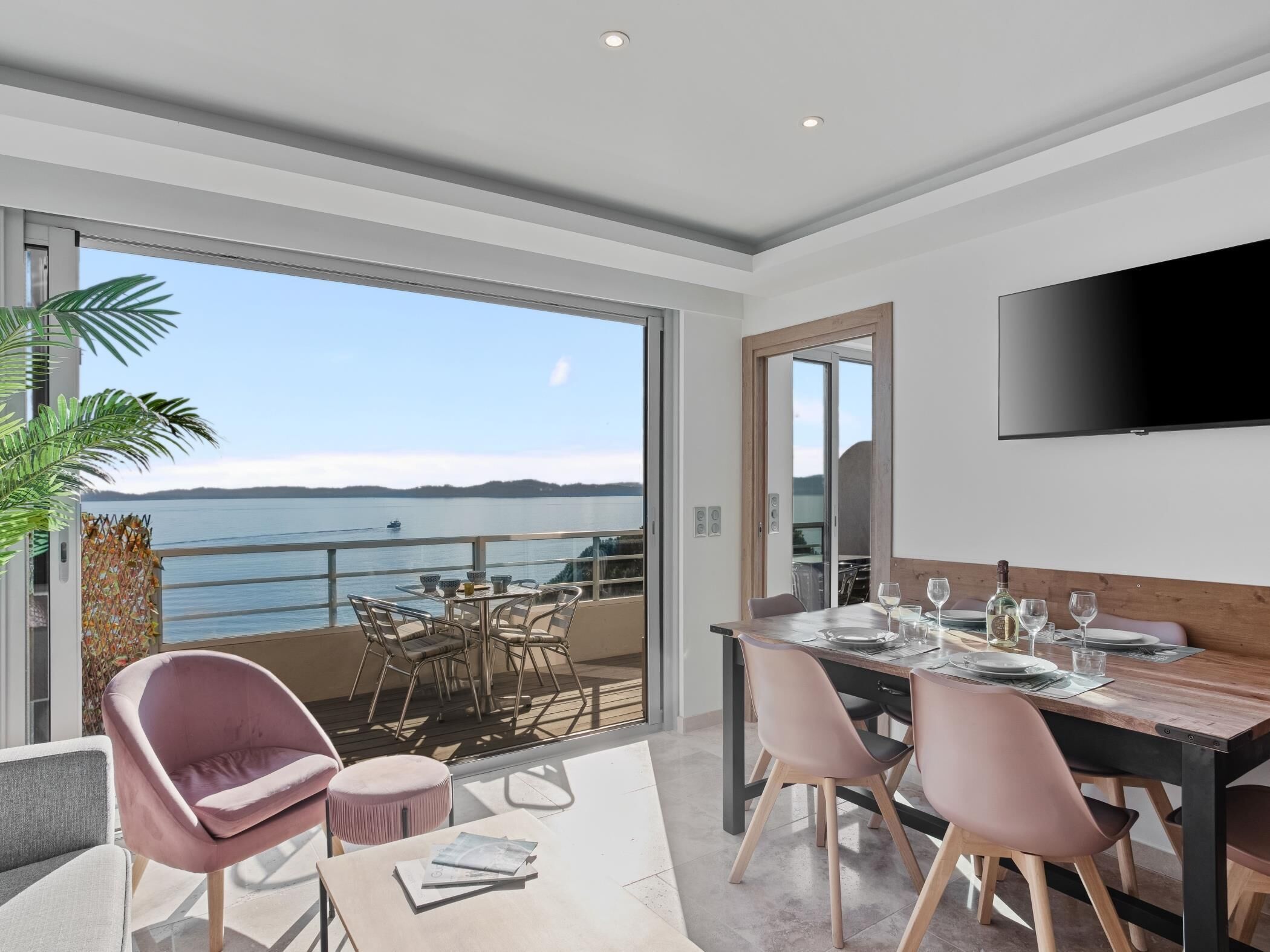 Een moderne eethoek met uitzicht op zee, een aan de muur bevestigde televisie en een balkon met buitenmeubilair.