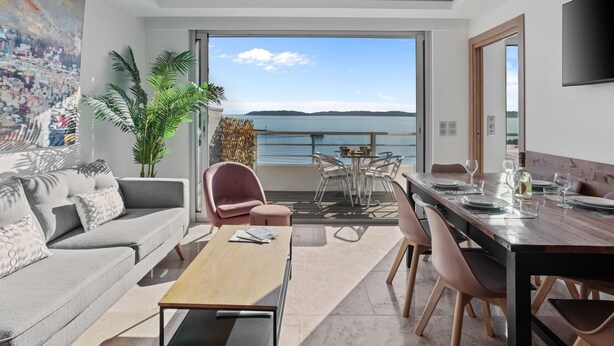 Una sala moderna con sofá, mesa de centro y comedor. Un ventanal ofrece vistas al mar.