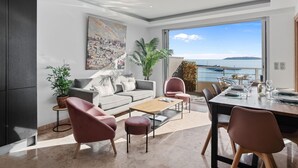 Una sala de estar moderna con sofá, mesa de centro y sillones, un comedor con mesa dispuesta para dos personas, y un amplio ventanal con vista al mar.