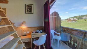 Een kamer met een houten bureau, stoelen, een bed, een raam met uitzicht op huizen en een lijst met een afbeelding aan de muur.