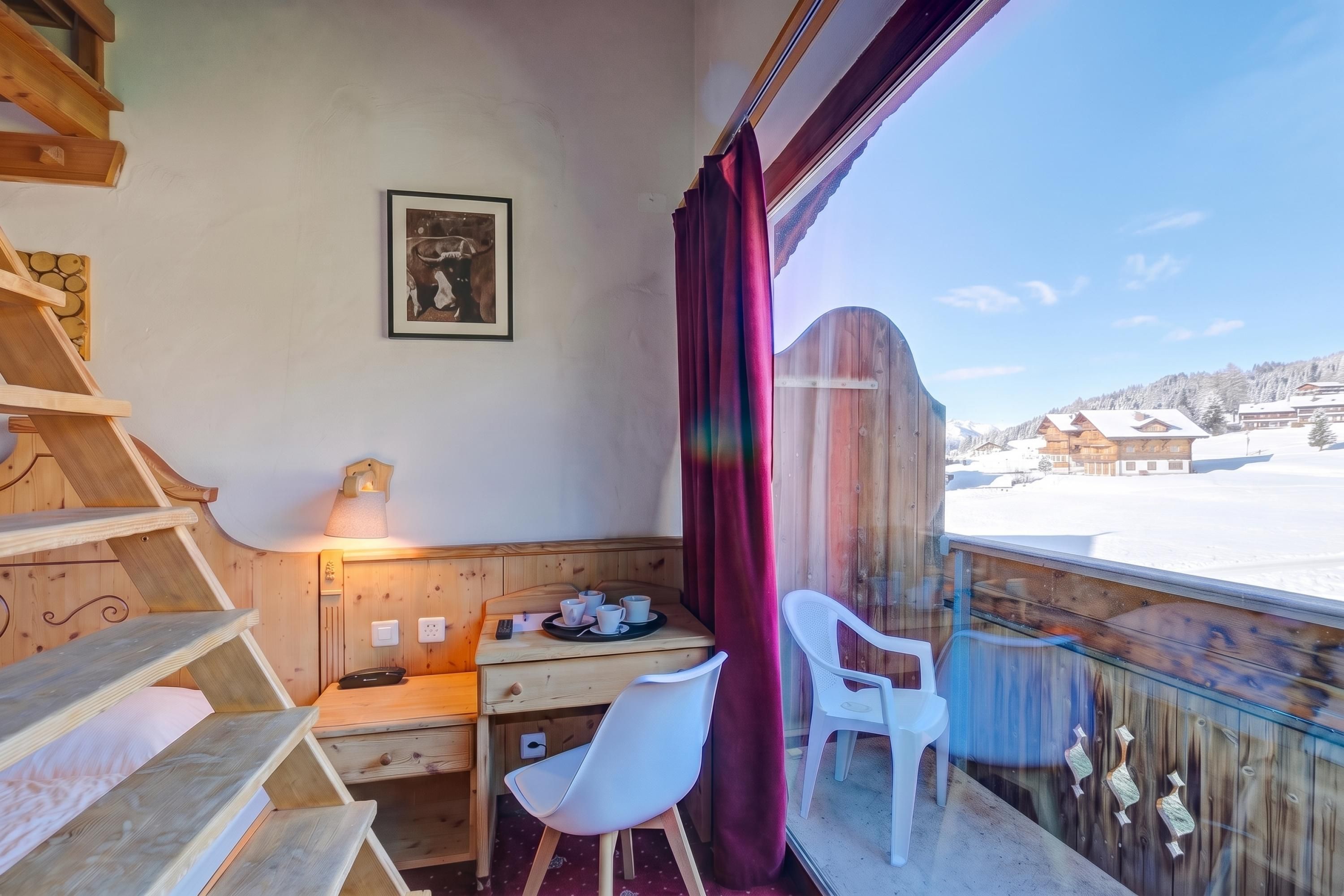 Una habitación acogedora con un escritorio de madera, una silla, una mesita con tazas, un cuadro en la pared y vistas a un paisaje nevado a través de una ventana con cortina.