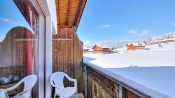 Ein hölzerner Balkon mit zwei weißen Plastikstühlen, der auf eine verschneite Landschaft mit mehreren Holzbauten blickt.
