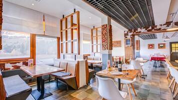 Ein Restaurant mit mehreren Essbereichen, darunter eine U-förmige Sitzgruppe und ein Bereich mit Tischen. Holzstühle und -tische sind vorhanden, und die Decke weist ein gitterartiges Muster auf.