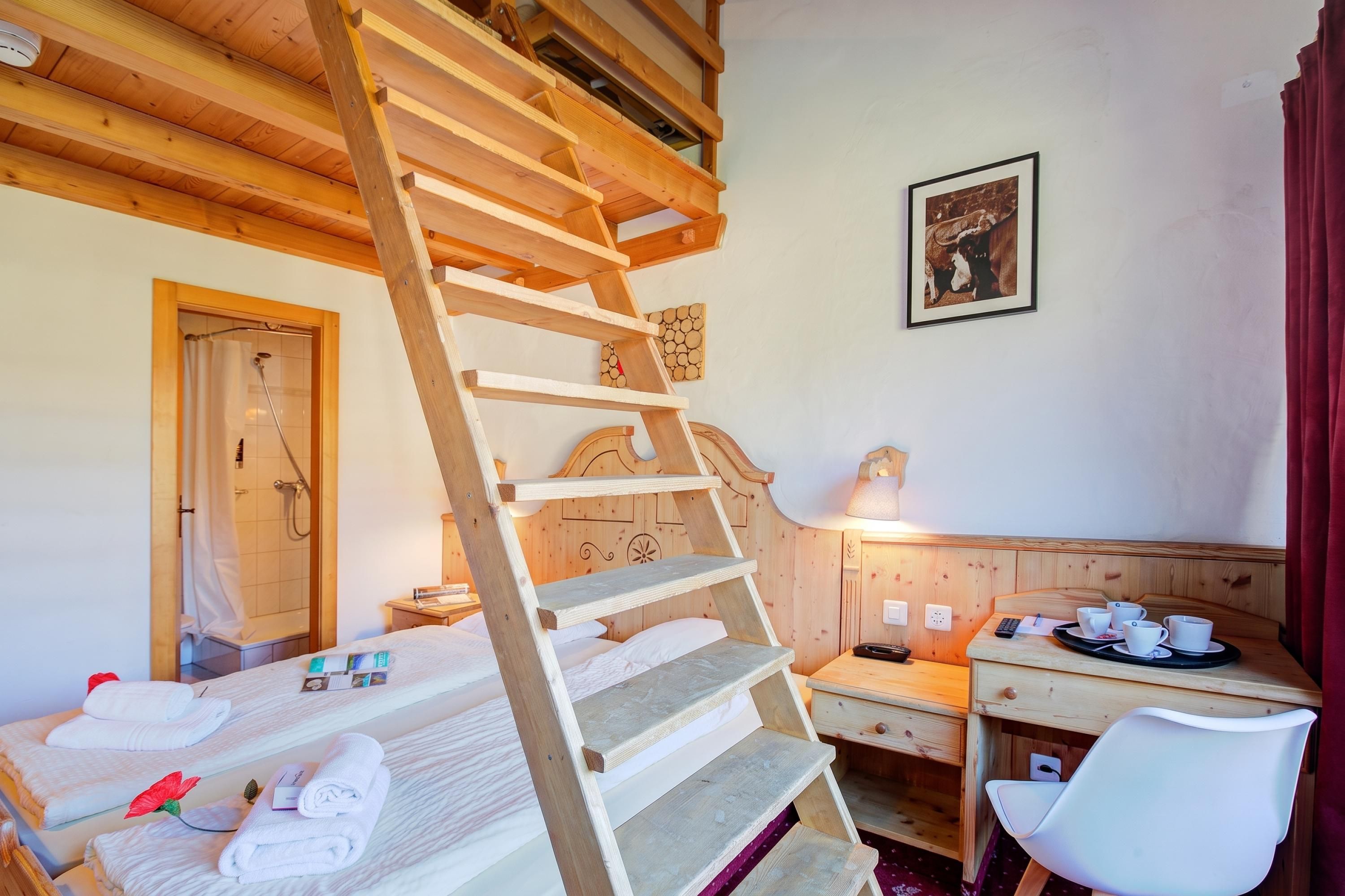A room with a wooden ladder leading to a bed, a desk with a lamp, and a framed picture on the wall.