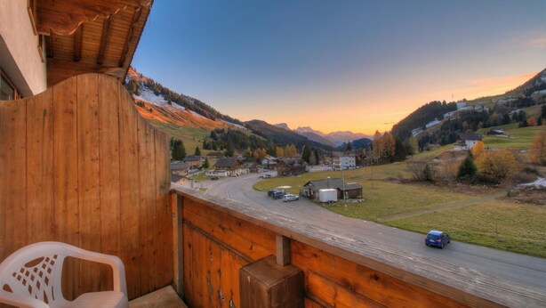 Un balcón de madera con vista a una calle con casas y montañas al fondo.
