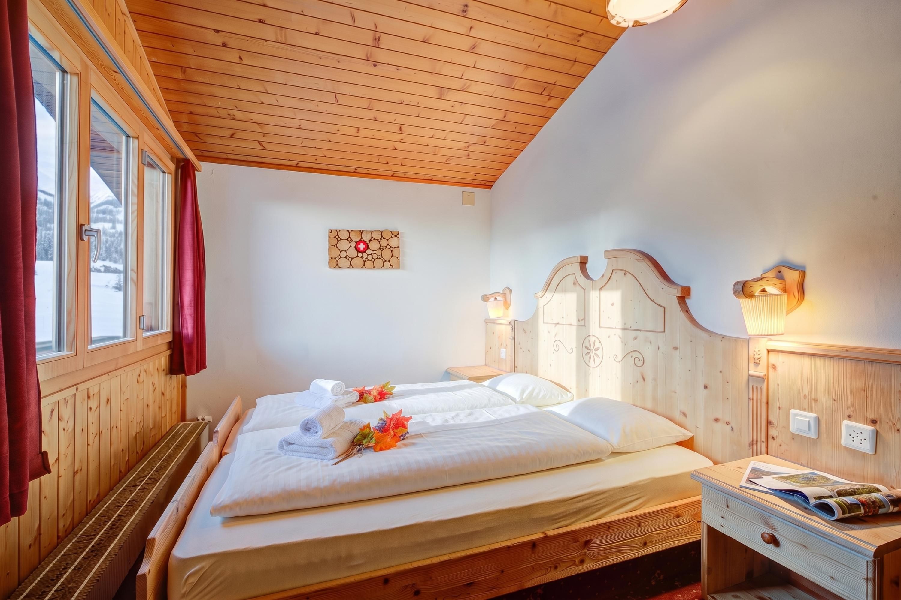 Una habitación de madera con una cama, un ventanal con cortinas, una lámpara de pared y una mesita con una revista.