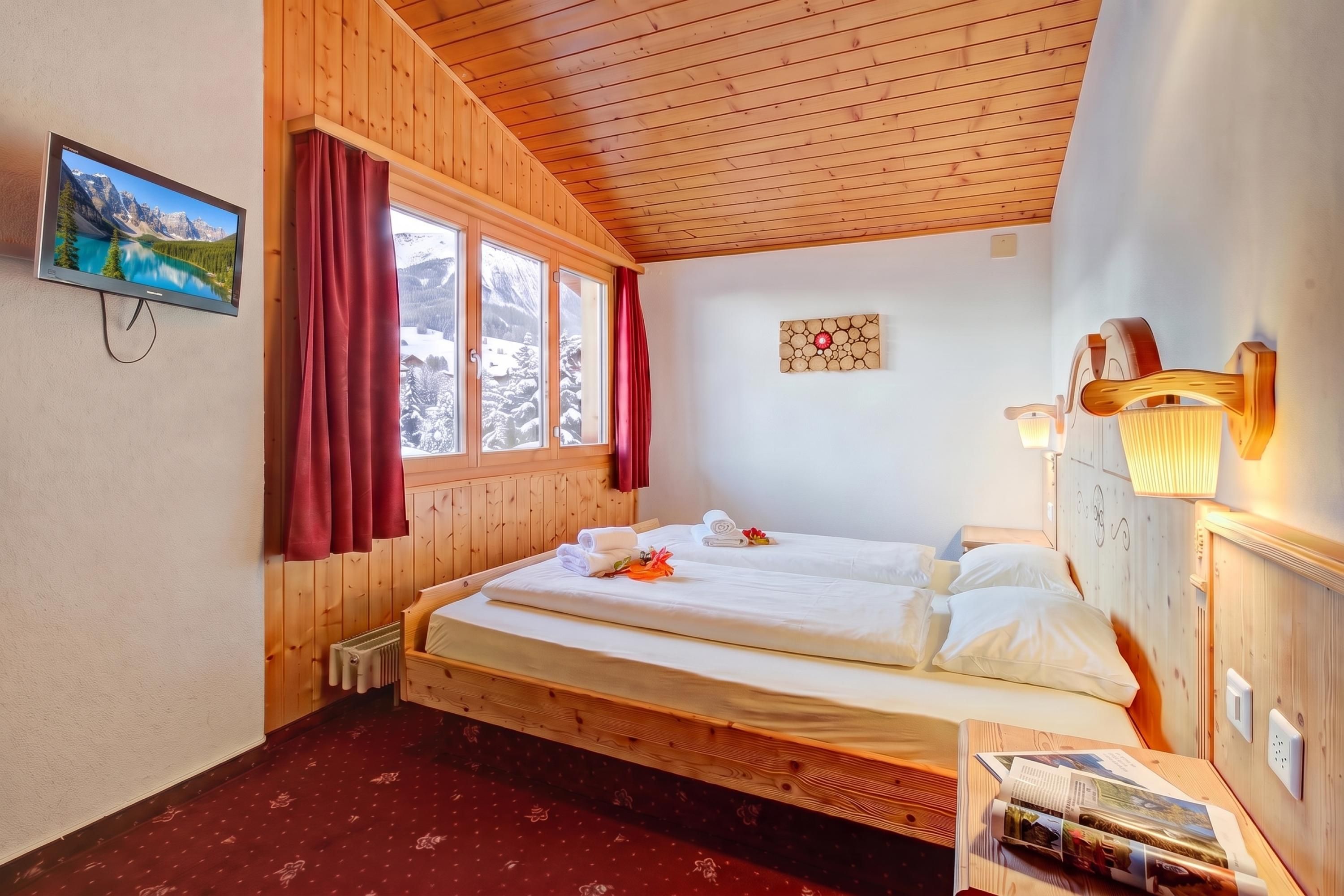 Ein Zimmer mit Holzwandverkleidung, einem Bett, einem Fenster mit Vorhängen, einer Wandleuchte und einem an der Wand befestigten Fernseher.