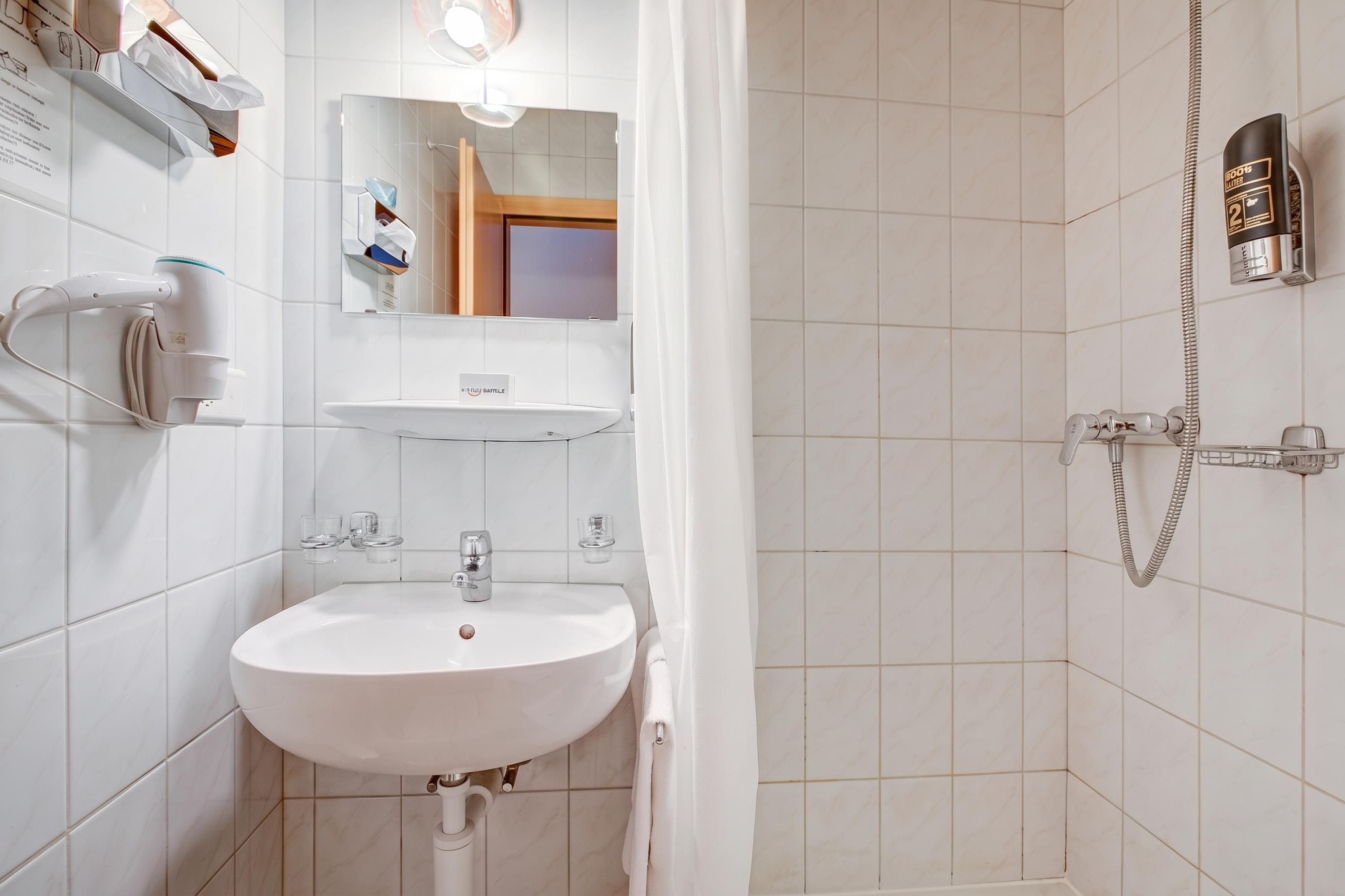 Ein Badezimmer mit einem weißen Waschbecken, einem Spiegel und einer Dusche. An der Wand befinden sich ein Handtuchhalter und eine Shampooflasche.