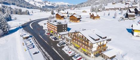 A snow-covered village with buildings, a winding road, and parked cars.