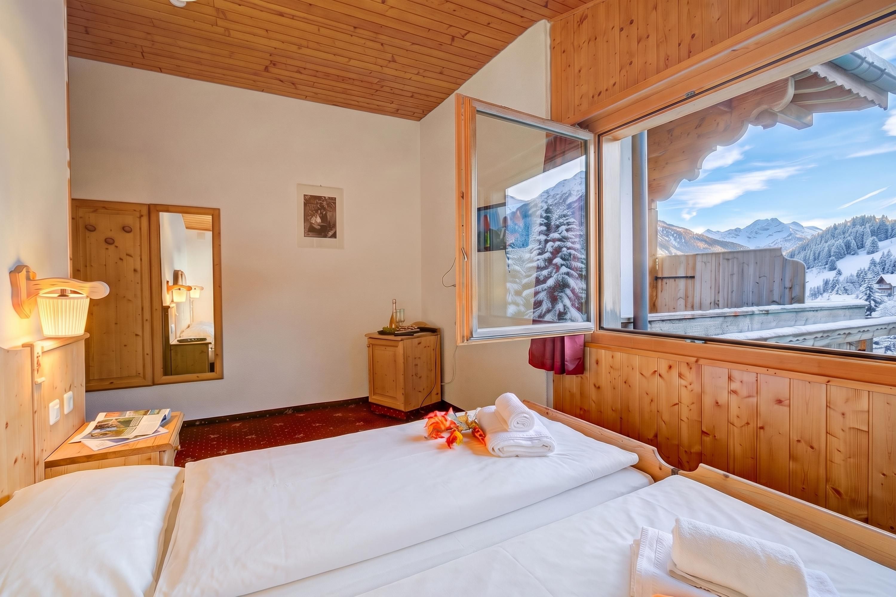 Ein Hotelzimmer mit Holzboden, einem großen Fenster mit Bergblick, einem Bett mit weißer Bettwäsche und einem kleinen Holzschrank.