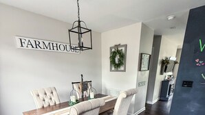 Ruokailutilassa on puinen pöytä, neljä verhoiltua tuolia, kattokruunu ja seinälle kiinnitetty kyltti, jossa lukee ”FARMHOUSE”.