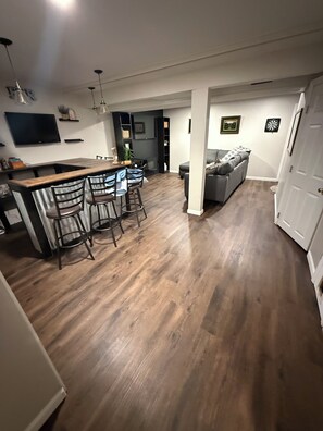 Uma sala de estar ampla com piso de madeira, um espaço de bar com banquetas, um sofá e uma televisão fixada na parede.
