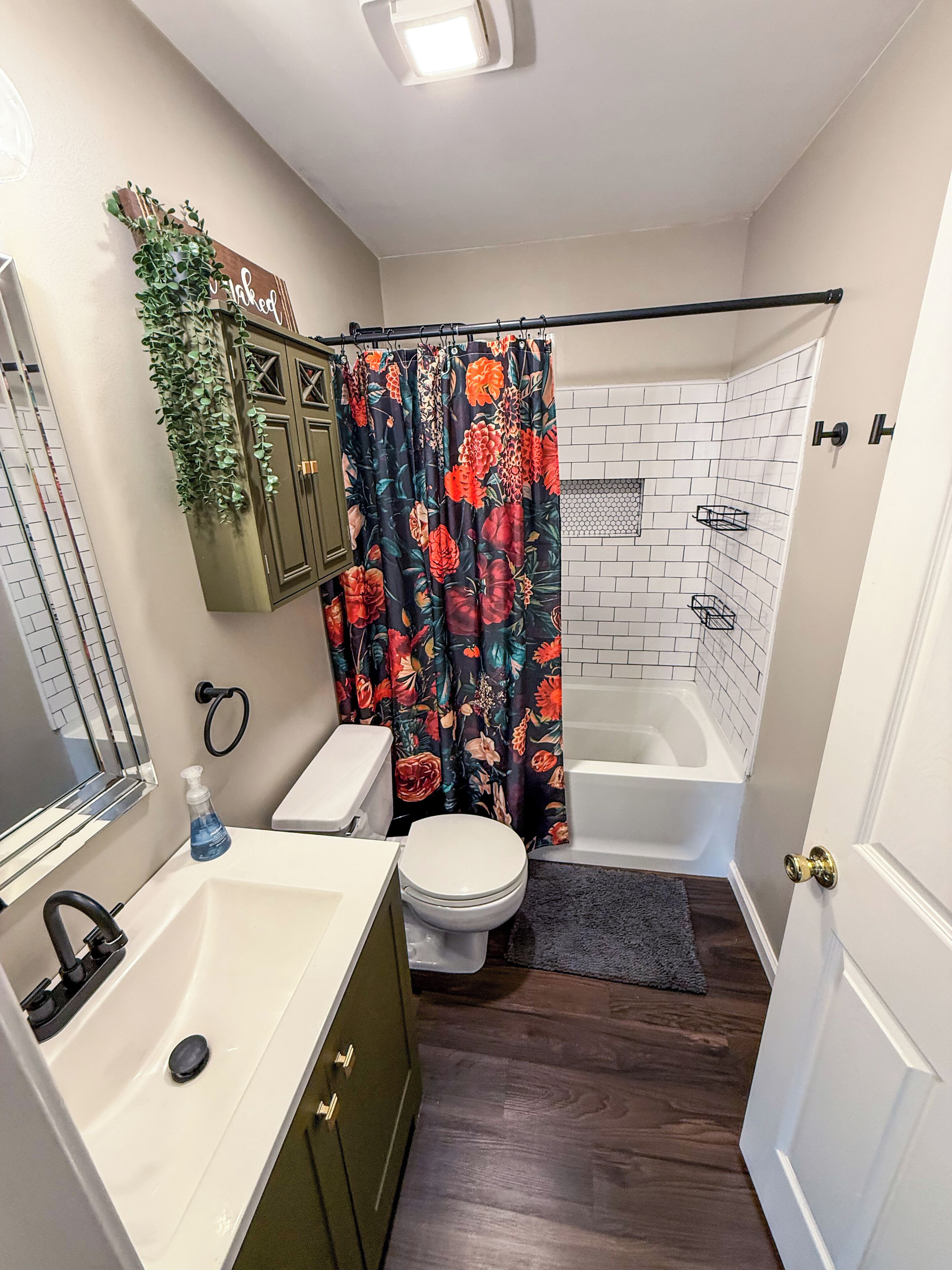 Un baño con lavamanos blanco, mueble verde, inodoro y ducha con cortina estampada. Hay un espejo sobre el lavamanos y un perchero en la pared.