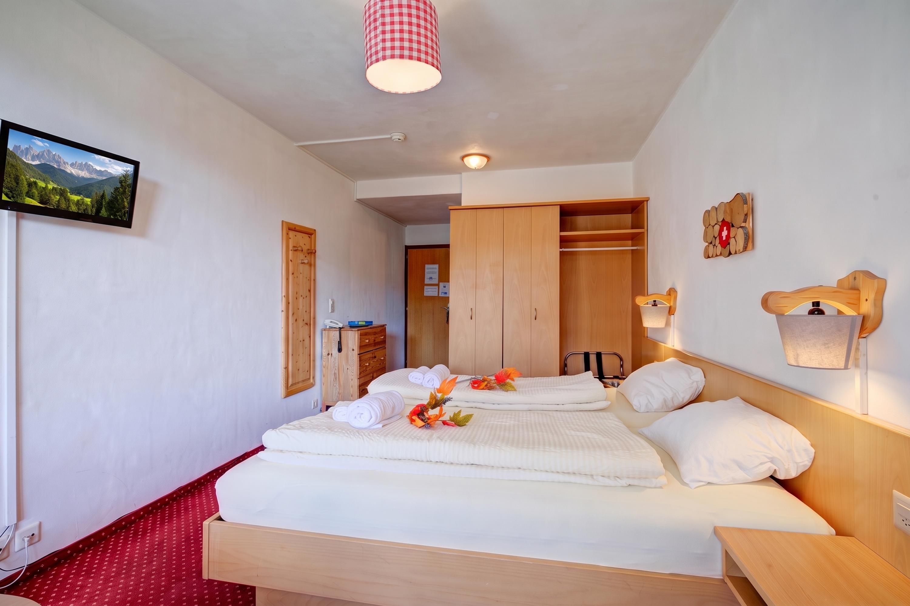 Ein Hotelzimmer mit einem Bett, einem Holzschrank, einem Schreibtisch, einem Telefon und einem an der Wand befestigten Fernseher.