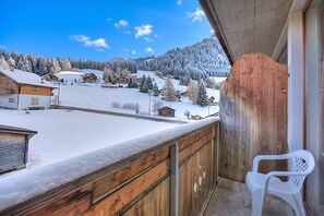 Un balcón con barandilla de madera, una silla de plástico blanca y un paisaje nevado con casas al fondo.