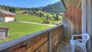Un balcón con barandilla de madera, una silla de plástico blanco y vistas a un valle verde con casas y montañas.