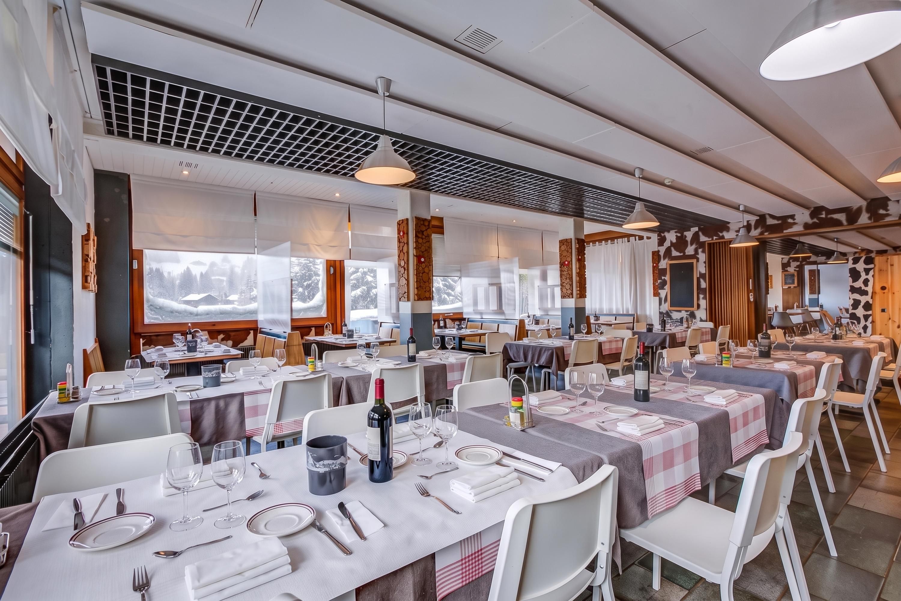 Ein Restaurant mit mehreren gedeckten Tischen, das modernes Interieur mit Pendelleuchten und einen Blick auf verschneite Berge durch große Fenster bietet.