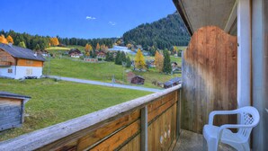 Un balcón con barandilla de madera, una silla de plástico blanco y vista a un pueblo con casas y una montaña al fondo.