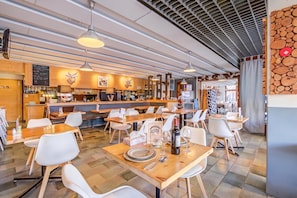 Interior de un restaurante con mesas de madera, sillas blancas, lámparas colgantes y una zona de bar con un tablero de menú.