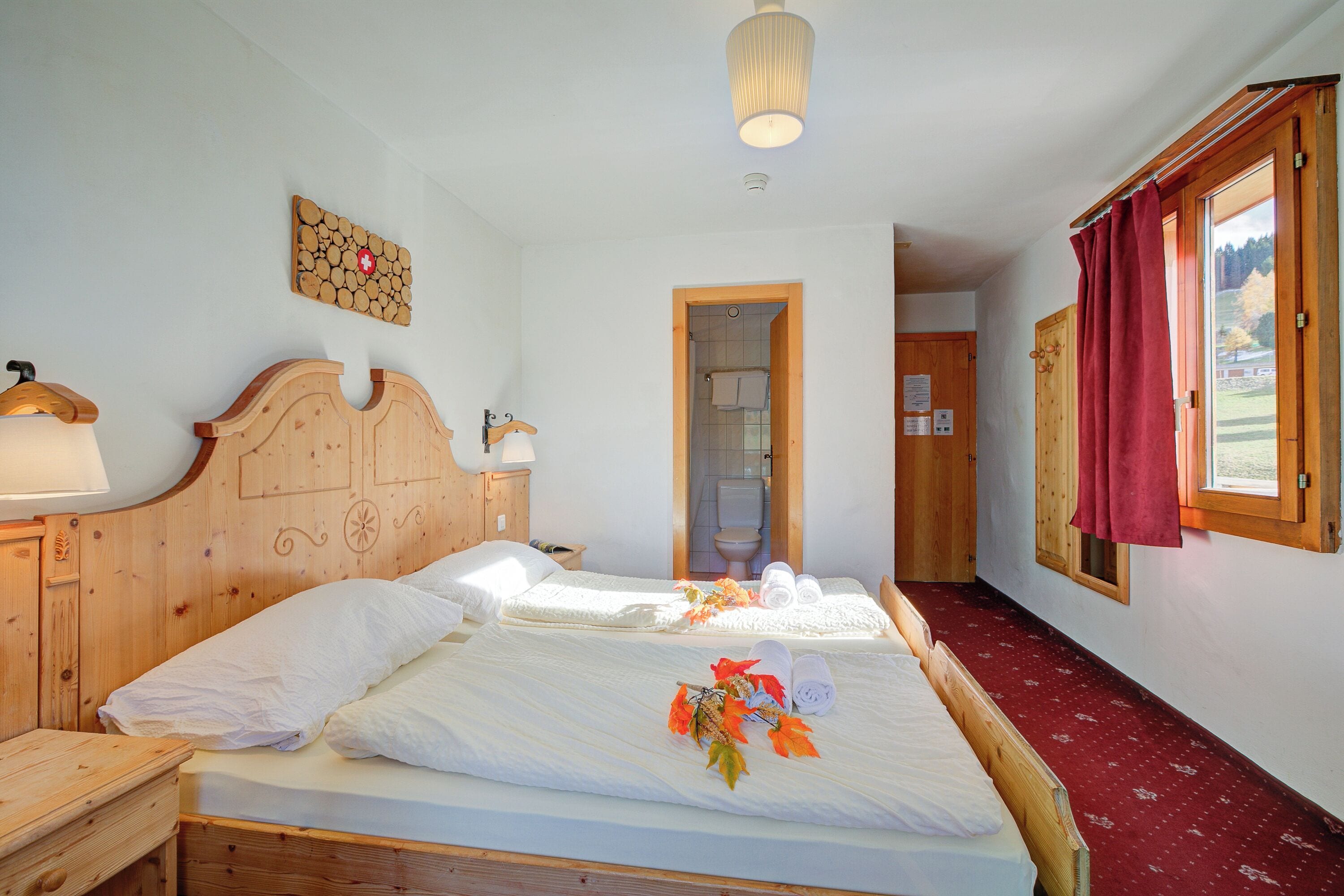 Ein Hotelzimmer mit zwei Holzbett, einem hölzernen Kopfteil, einem Nachttisch aus Holz und einer Wandlampe. Ein Fenster mit rotem Vorhang.