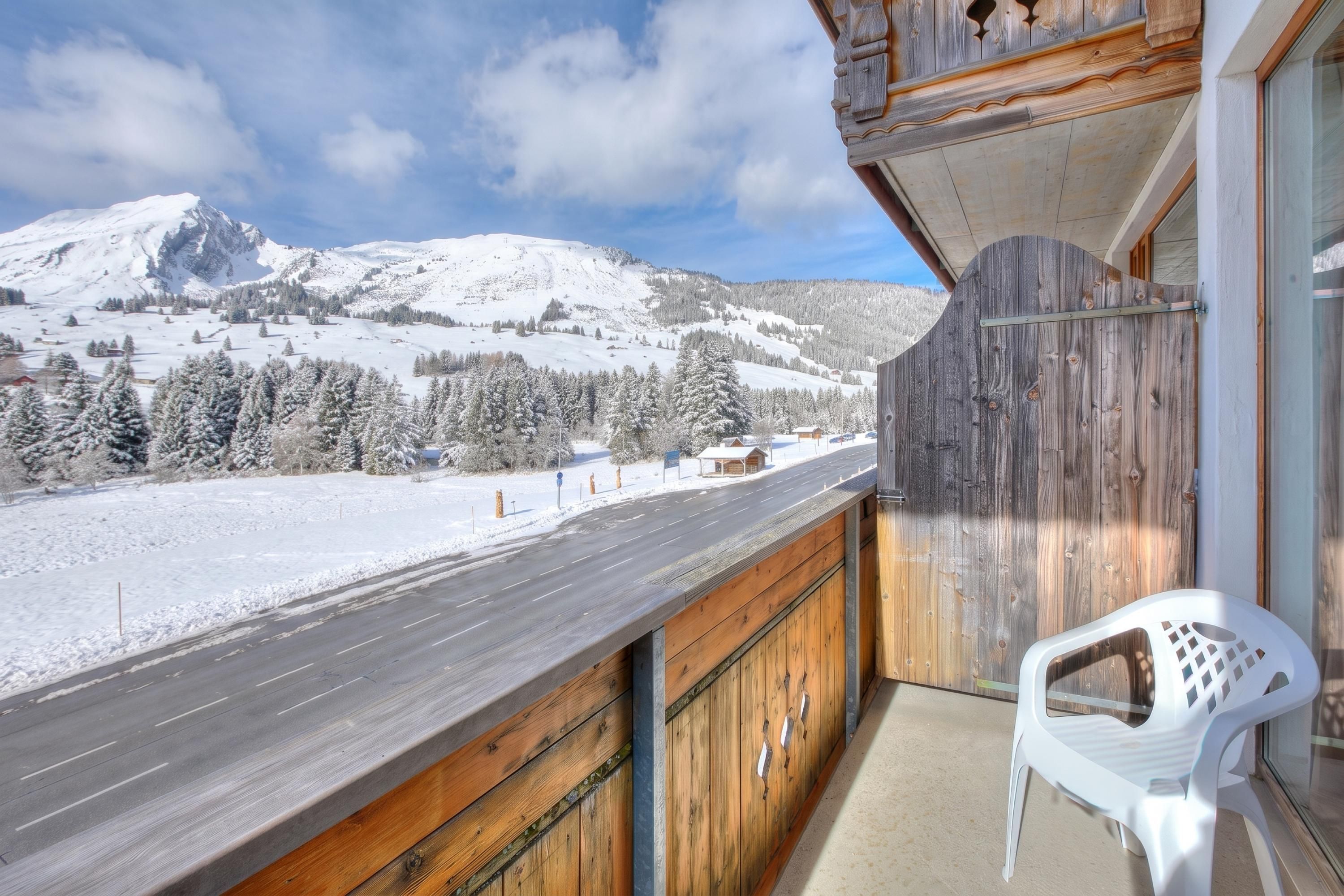 Un balcon donnant sur un paysage montagneux enneigé, avec une balustrade en bois et une chaise en plastique blanche.