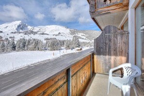 Un balcón con vistas a un paisaje montañoso nevado, barandilla de madera y una silla de plástico blanco.