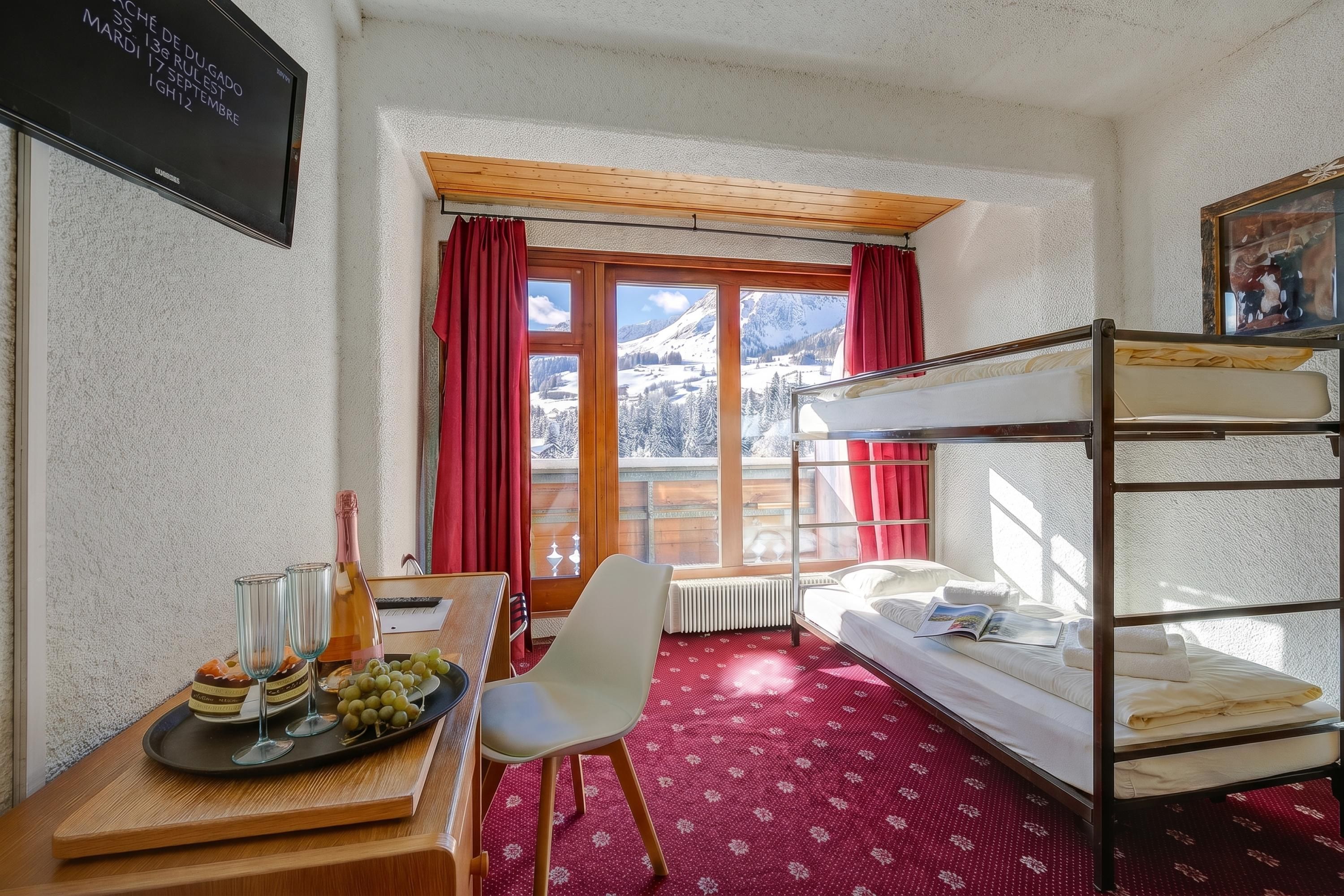 Una stanza con un letto a castello, una scrivania con una bottiglia e dei bicchieri, una sedia e una finestra vista montagne innevate.