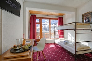 Una habitación con literas, un escritorio con una botella y vasos, una silla y una ventana con vistas a montañas nevadas.