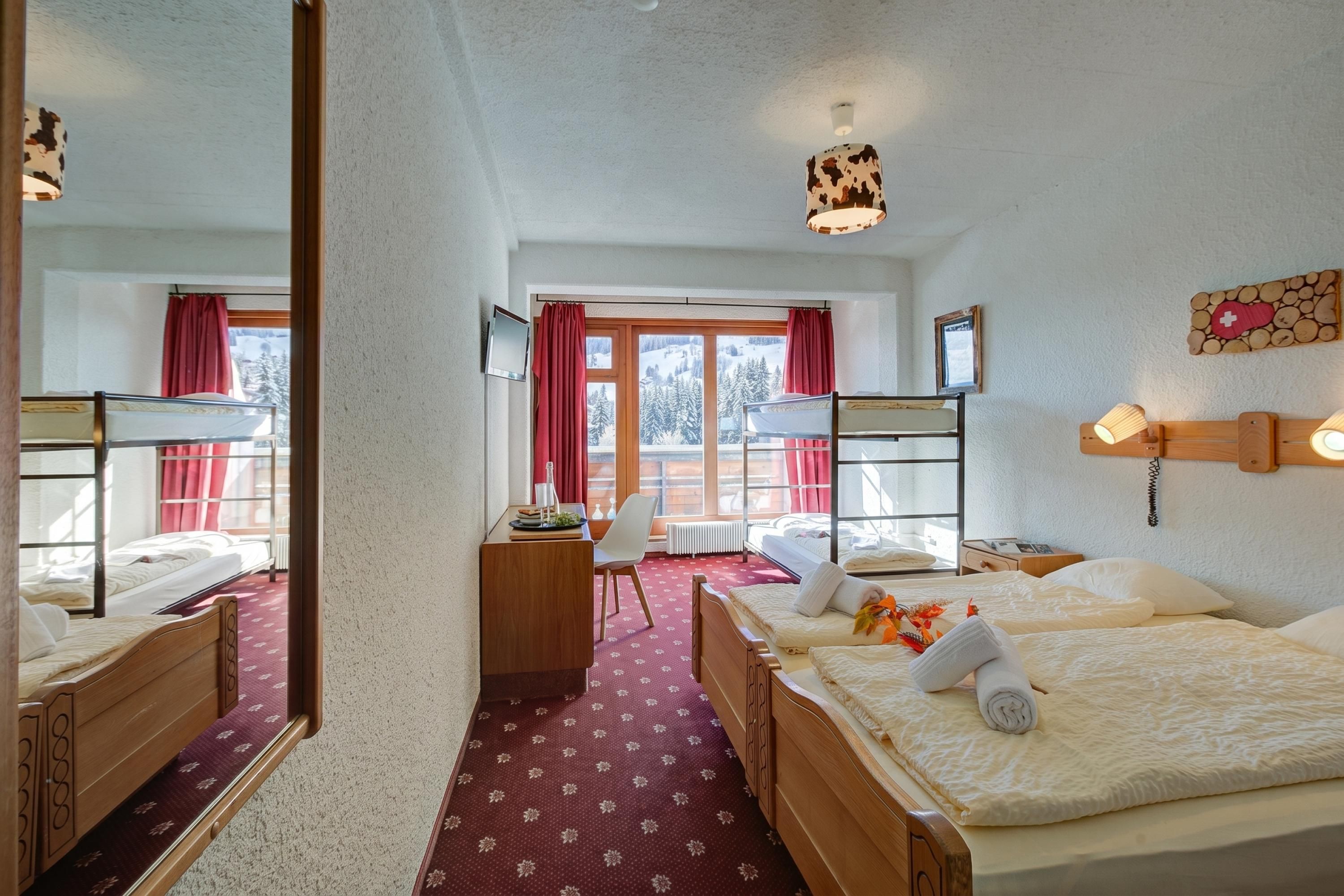 A hotel room with bunk beds, a desk, a chair, a window with curtains, and a view of a snowy landscape.
