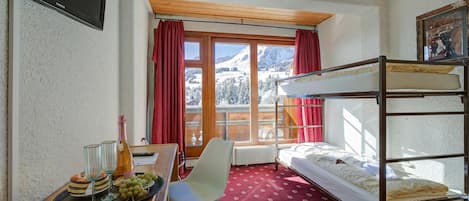 Habitación con literas, una mesa con una botella y vasos, una silla y una ventana con vista a montañas.