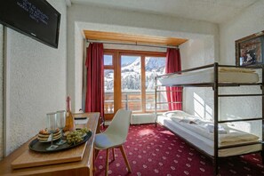 Una habitación con literas, un escritorio con una botella y vasos, una silla y una ventana con vista a montañas.