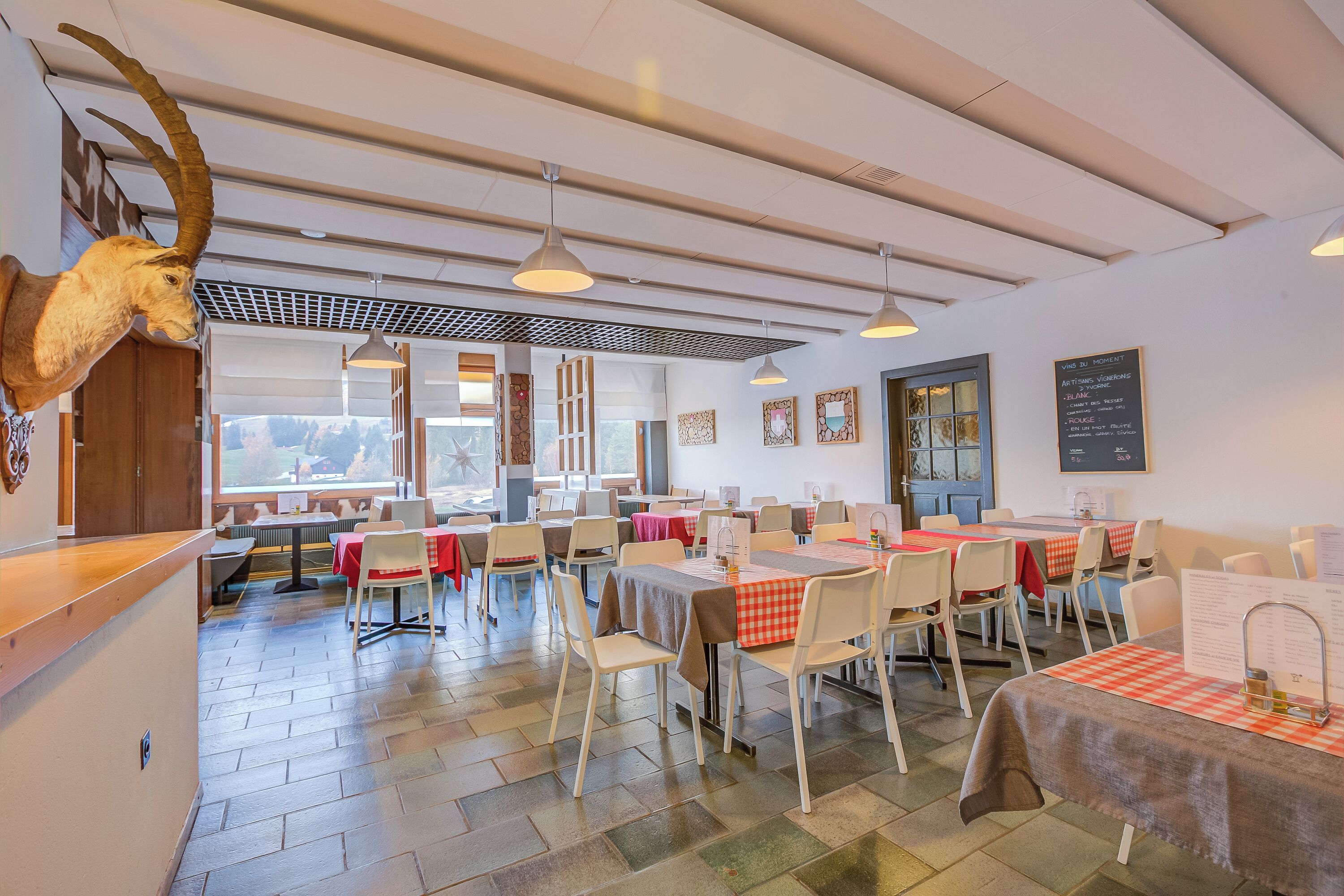 Un restaurante con techos altos, iluminación colgante y una cabeza de animal montada en la pared. Las mesas están cubiertas con manteles a cuadros rojo y blanco, y sillas dispuestas alrededor.