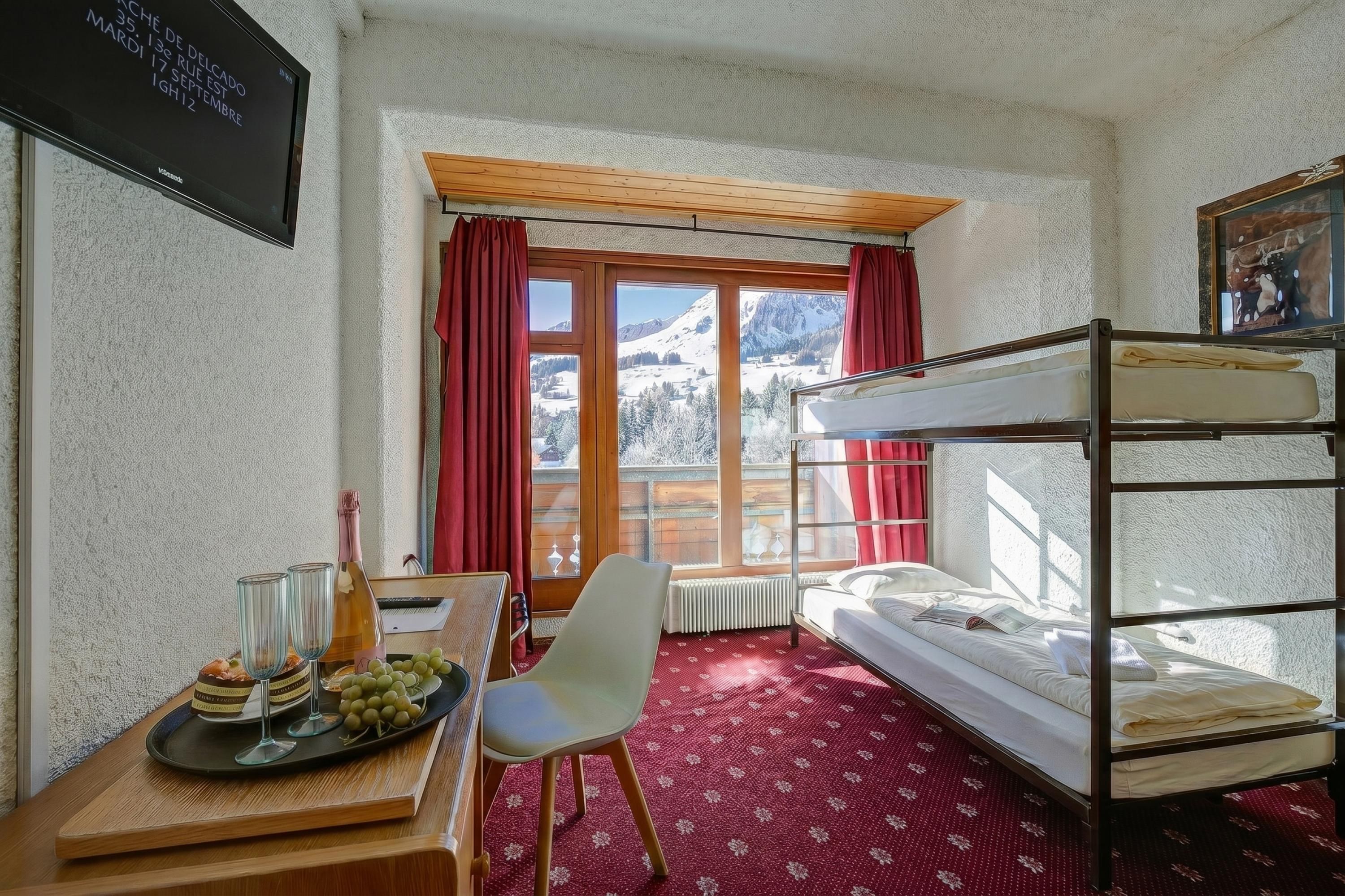 Une chambre avec un lit superposé, un bureau avec une bouteille et des verres, une chaise et une fenêtre donnant sur les montagnes.