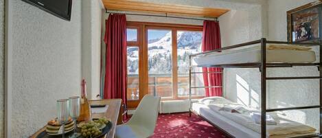 A room with a bunk bed, a desk with a bottle and glasses, a chair, and a window with a view of mountains.