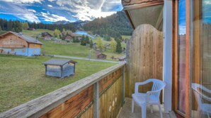 Un balcon en bois offrant une vue sur un village, une petite construction en bois et une chaise en plastique blanche.