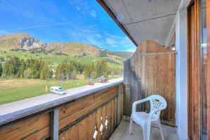 Un balcon donnant sur un paysage montagneux, une chaise en plastique blanche et une rambarde en bois.