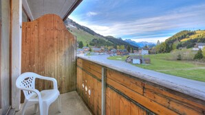 A balcony with a wooden railing, a white plastic chair, and a view of a town with houses and mountains in the distance.