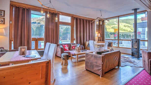 A cozy living room with a wooden table, sofa, and chairs, large windows with curtains, and a fireplace.