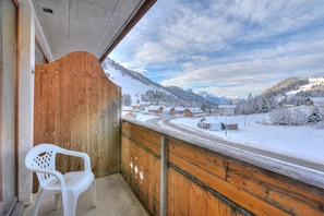 A wooden balcony with a white plastic chair, overlooking a snow-covered village and mountains.