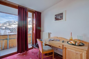 A hotel room with a wooden desk, a chair, a bed, a window with curtains, a radiator, a framed picture on the wall, and a view of snow-covered mountains.