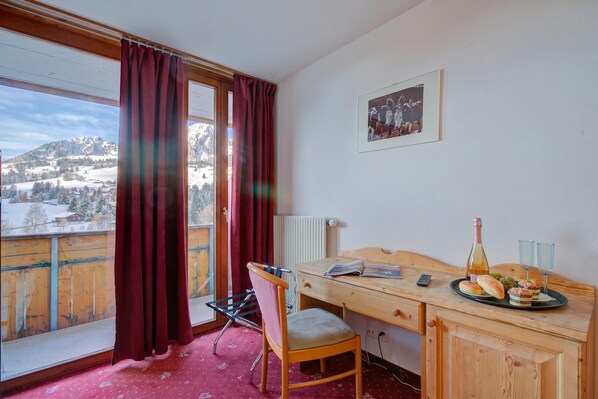 Habitación de hotel con un escritorio de madera, una silla, una cama, un ventanal con cortinas, un radiador, un cuadro en la pared y vistas a montañas nevadas.