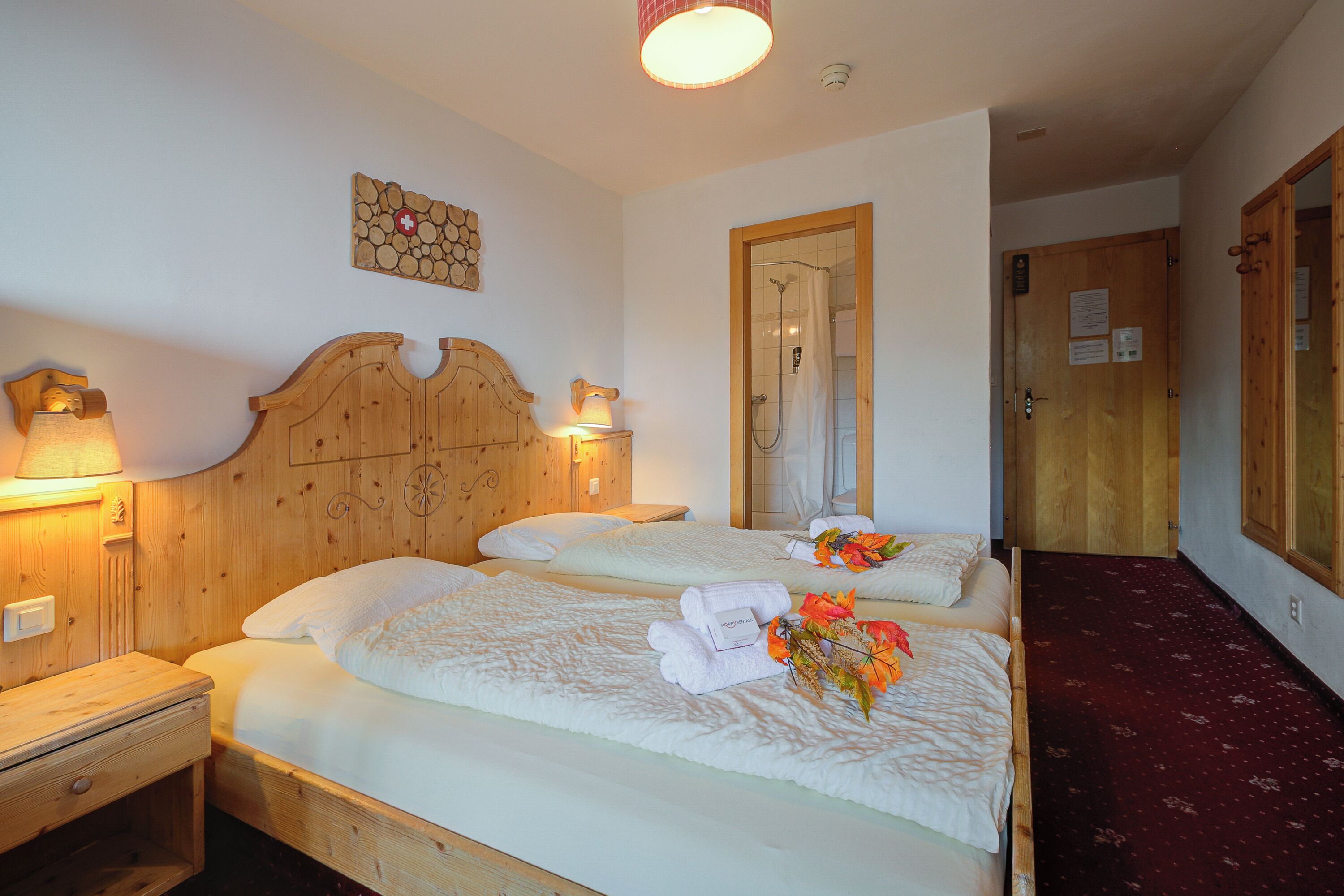Une chambre d’hôtel avec deux lits en bois, une tête de lit en bois, une table de chevet en bois, une lampe fixée au mur et une porte en bois.